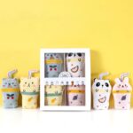 Box of kawaii plush catnip drink toys surrounded by loose toys featuring cat, panda, rabbit, and bear designs on a yellow background.