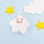 Close up of white catnip monster plush toy with a circle highlighting dried catnip filling, with felt foliage and a purple toy in the background against a yellow background.